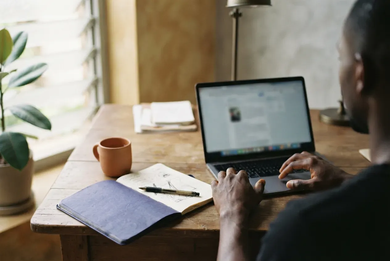 A man writing on a laptop at a wooden desk, beside an open notebook and a mug