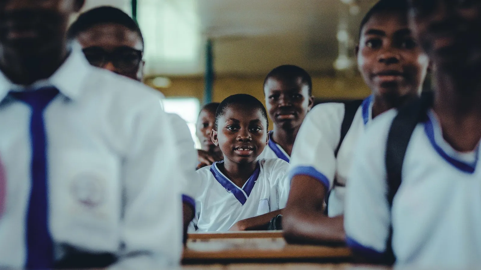 Secondary school students in white and blue uniforms fill a classroom, one looking towards the camera between the others.