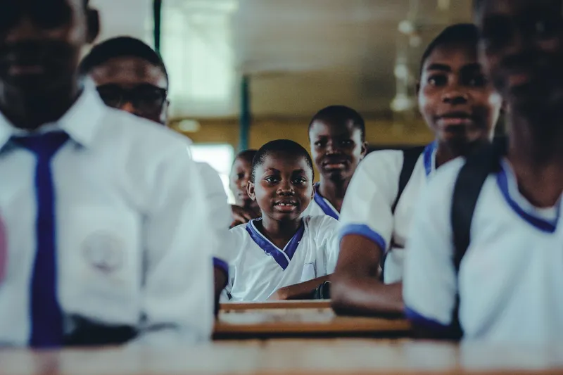 Secondary school students in white and blue uniforms fill a classroom, one looking towards the camera between the others.
