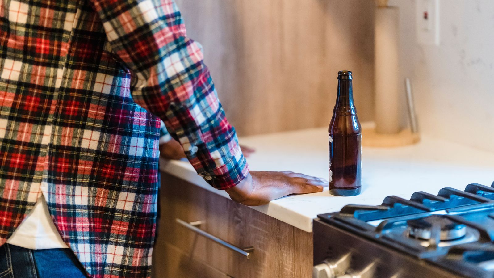 concept shot of a man in a kitchen with an open bottle of beer. Both hands are on the kitchen counter and the beer is beside him. Only his back and side are visible but his posture depicts contemplation