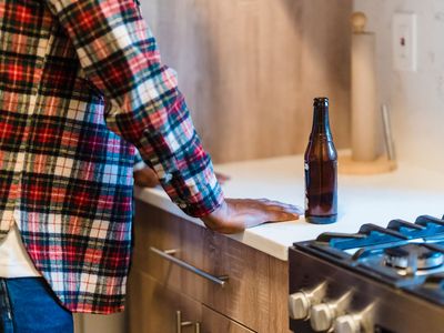 concept shot of a man in a kitchen with an open bottle of beer. Both hands are on the kitchen counter and the beer is beside him. Only his back and side are visible but his posture depicts contemplation