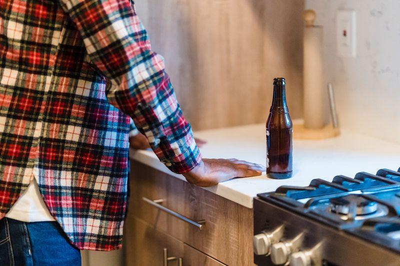 concept shot of a man in a kitchen with an open bottle of beer. Both hands are on the kitchen counter and the beer is beside him. Only his back and side are visible but his posture depicts contemplation