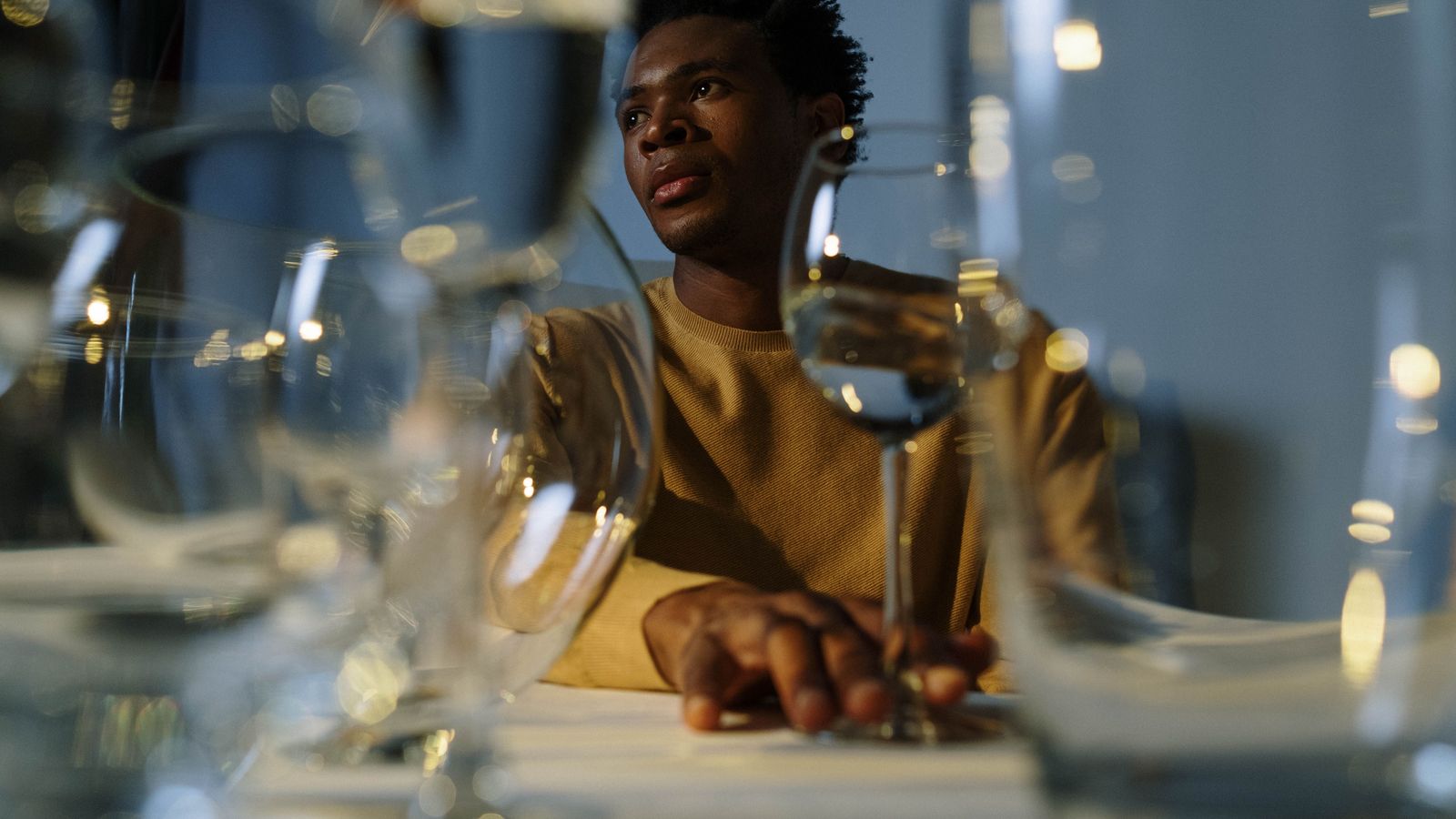 A young man sits at a table of empty wine glasses, one hand resting on a stem, looking away from them.